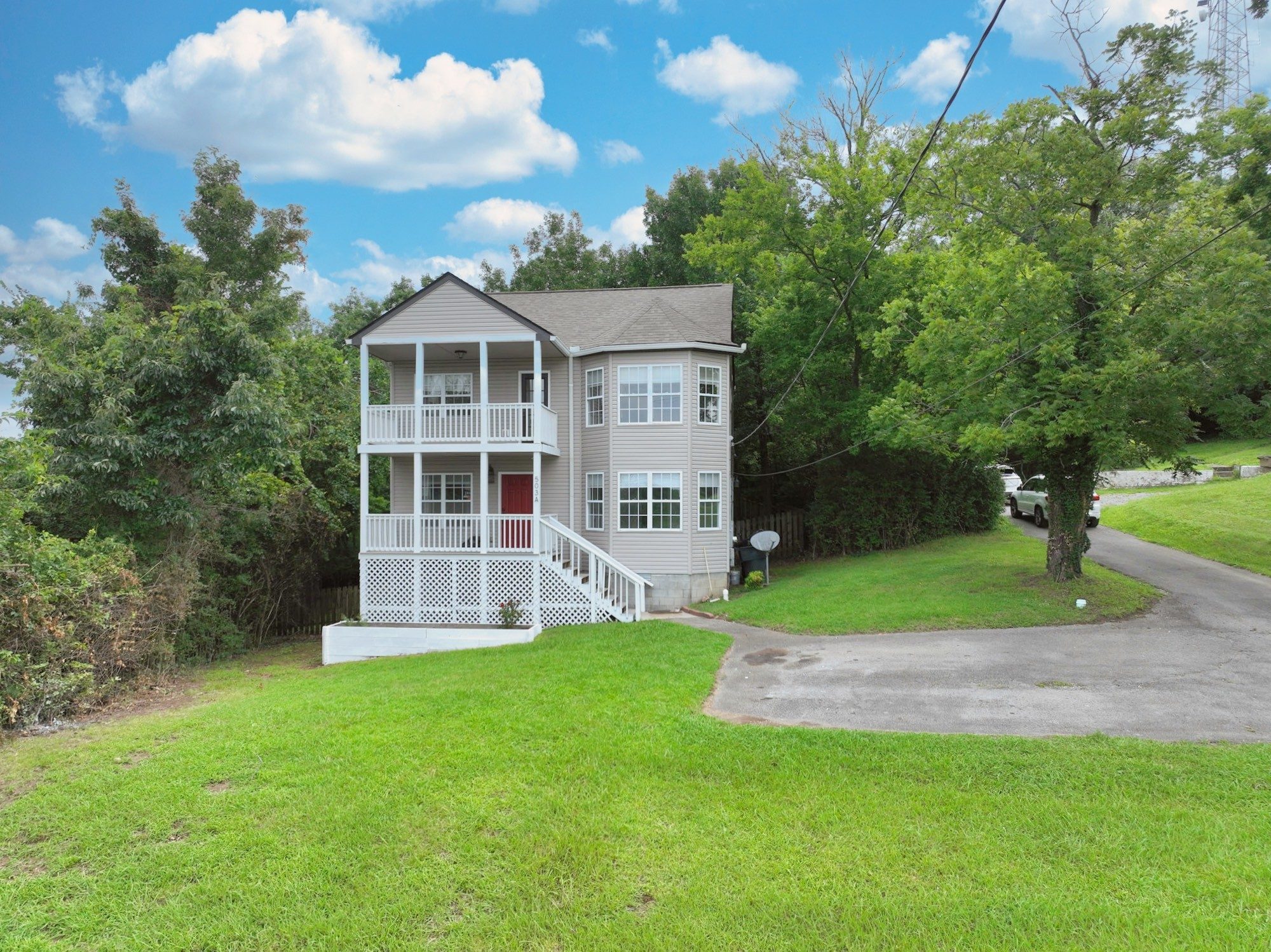 Front exterior of 503 Ben Allen Road, a two story home with light siding, a covered front porch with white railings, a matching second floor deck, and a long driveway on a wooded lot, one of the East Nashville homes for sale in Nashville